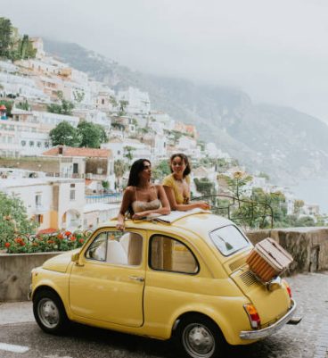 Two beautiful young woman inside a small, vintage yellow car. The top remains open. They stand up in the vehicle, through the sunroof and admire the view as they talk to one another. We can see Italy's famous view of Positano in the background. Depicts a scene of experiential travel, suggesting renting a car, travel tours, European road trips and nostalgia.